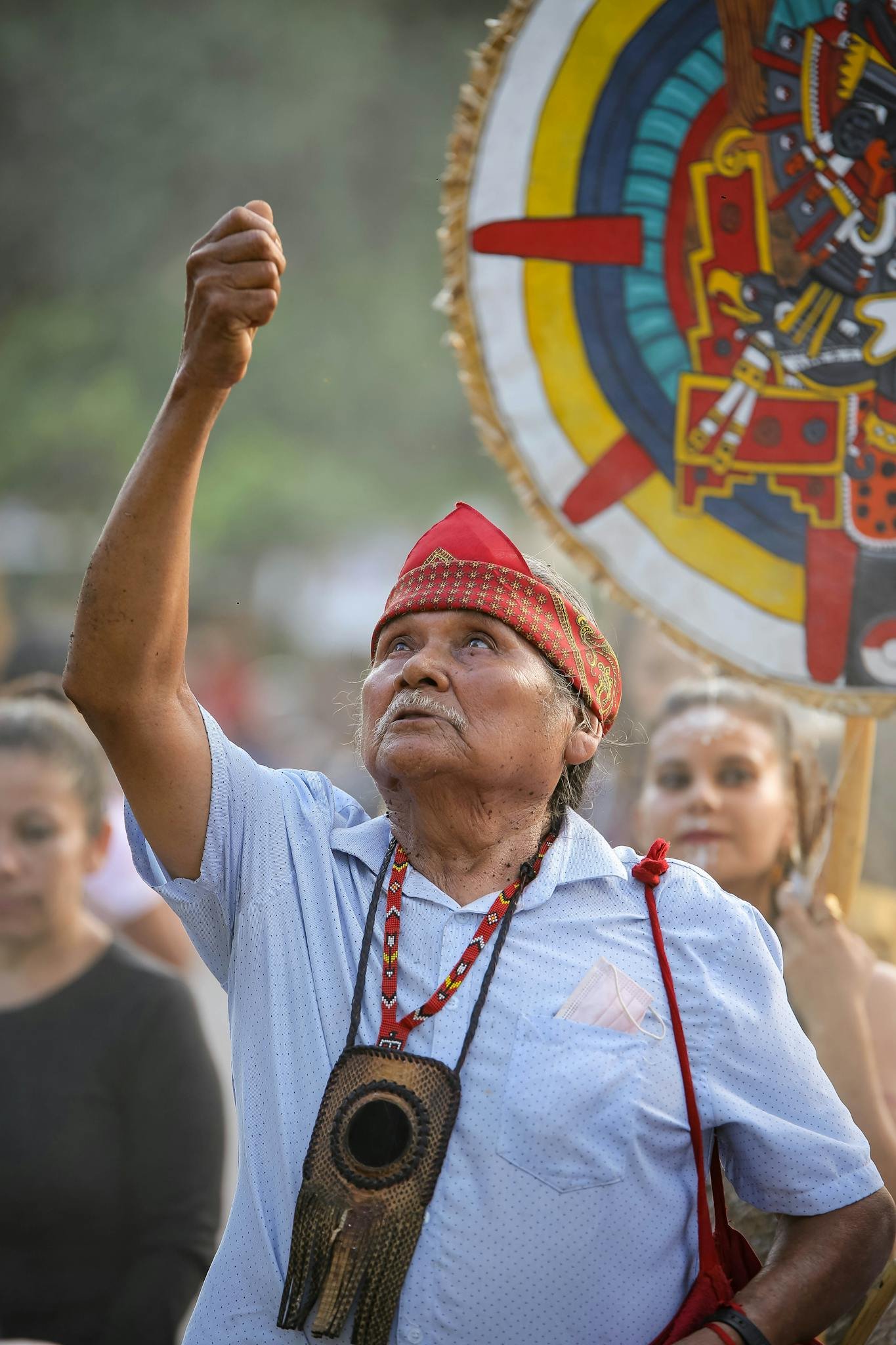 Elderly man in traditional attire participates in a cultural ceremony, holding up his hand outdoors.