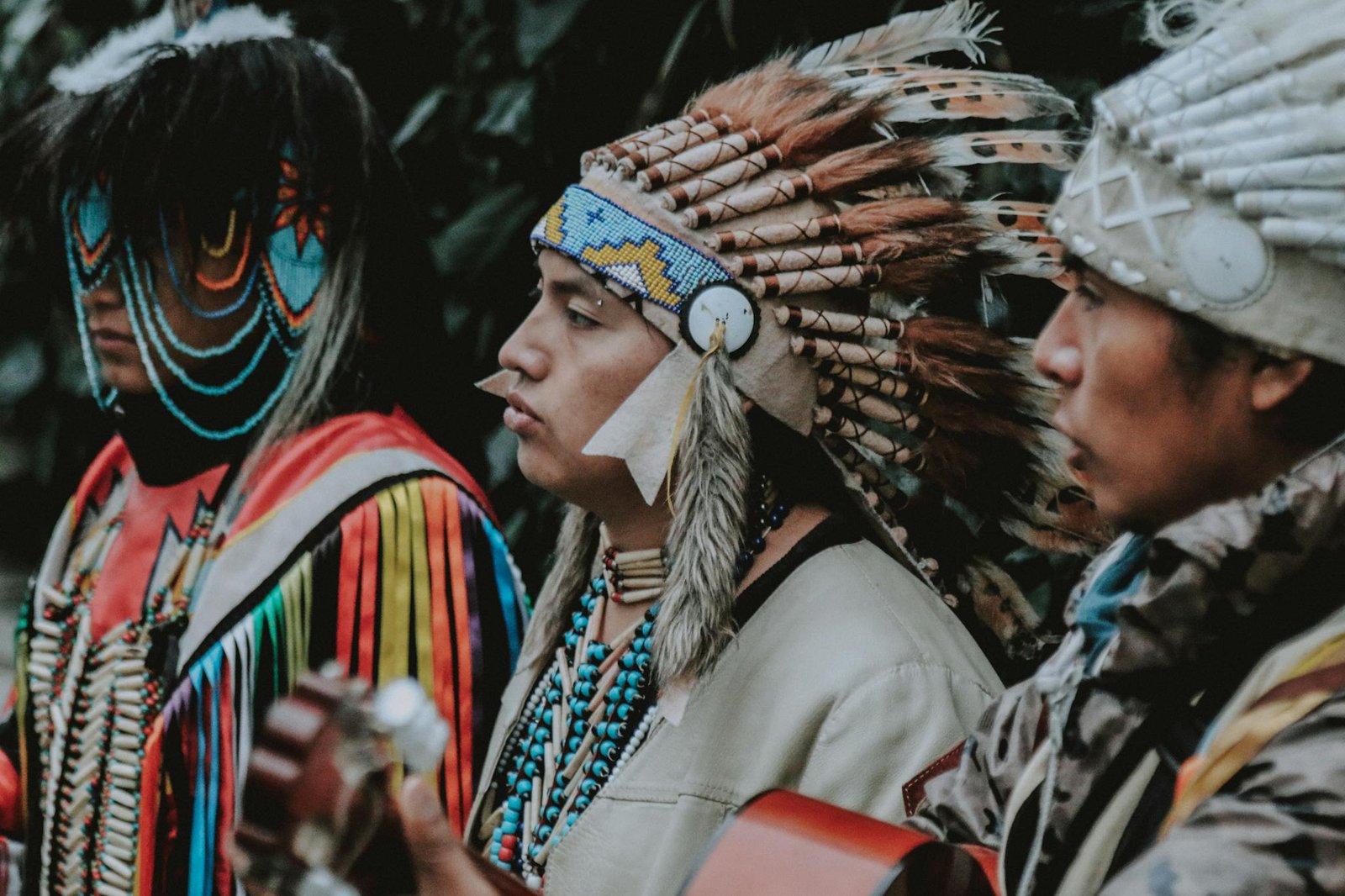 Three Native American men in traditional attire play guitars outdoors, capturing a cultural musical moment.